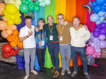 Five smiling people stand arm-in-arm posing for a photo in front of a rainbow-colored fabric backdrop flanked by vibrant balloon arches and mini disco balls.