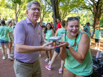 A smiling man wearing glasses and a patterned short-sleeved shirt hands a small, clear container filled with colorful items to an excited student in a light green top outdoor on a brick walkway. Other students wearing light green shirts are gathering and socializing in the background surrounded by shade trees.