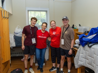 Four smiling people—two students wearing orientation lanyards and two women wearing red Davidson College shirts—pose together for a photo inside a college dorm room filled with move-in bags and dorm furniture.
