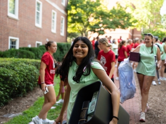 A smiling student in a light green top carries a black storage bin filled with supplies along a brick walkway, surrounded by other students carrying move-in belongings in front of a brick dorm building.