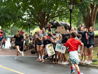A young boy in a red shirt runs along a paved road past a crowd of cheering spectators holding handmade encouragement signs.