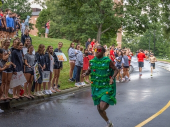 A runner wearing a green Christmas tree costume and red sunglasses races down a wet road past cheering spectators holding signs.