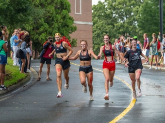 Four female runners wearing costumes, face paint, and athletic gear hold hands while sprinting down a wet campus road.
