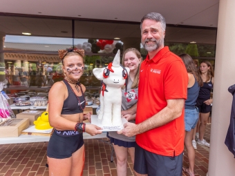 A man in a red Davidson Athletics polo shirt and two students hold a large, sculpted white unicorn cake together outdoors.