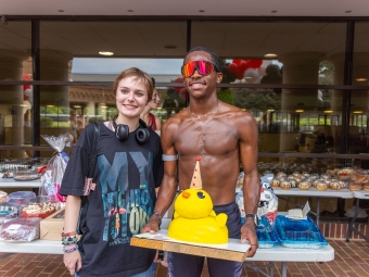 A smiling runner in red sunglasses holds a yellow duck-shaped cake alongside a friend in front of tables filled with baked goods.