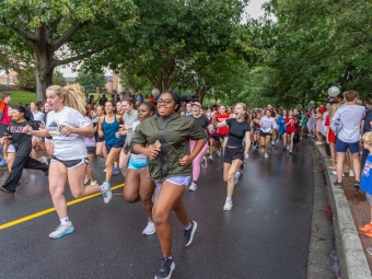 A large group of smiling young runners race down a tree-lined paved road while spectators cheer along the sidewalk.