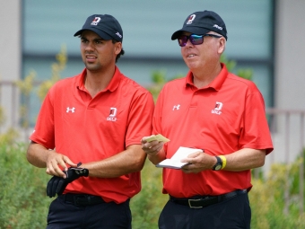 A young golfer adjusting a black glove next to a coach reviewing notes on a small yardage book. Both are wearing matching red Under Armour polo shirts and black caps branded with a white "D" and "GOLF" logo.