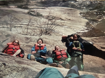 Five people wearing life jackets and outdoor gear rest on a large, flat rock outcrop near a running river or stream.