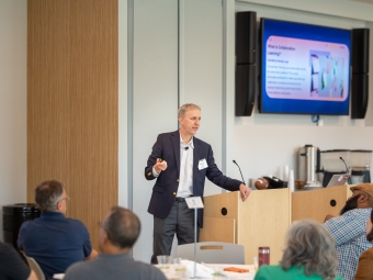 A blonde man stands at a podium in front of a TV screen, addressing a seated audience
