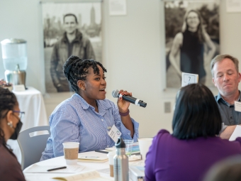 A woman with braids and a blue shirt holds a microphone, seated