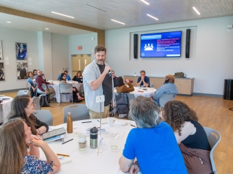 A man stands and speaks in a room full of adults seated at round tables