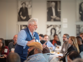 An older man in a blue vest stands and speaks in a room full of people