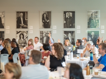 A room with portraits hung on the wall. Adults with hands raised, seated at round tables