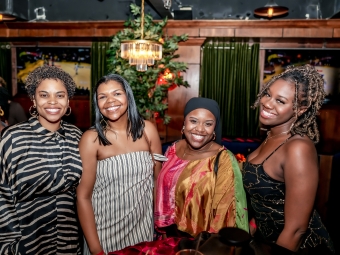 Four smiling women pose together side-by-side in a dimly lit lounge with wooden paneling.
