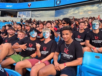 Students seated in the stands of the soccer stadium in Charlotte, with blue facepaint and Davidson College shirts