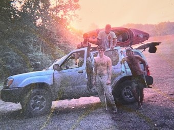 Four people pose around an SUV loaded with a kayak on its roof rack at sunset/sunrise.