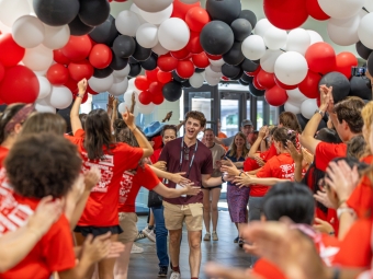 A young man walking down a hallway filled with red, white, and black balloons while crowd members in matching red shirts cheer and high-five him.