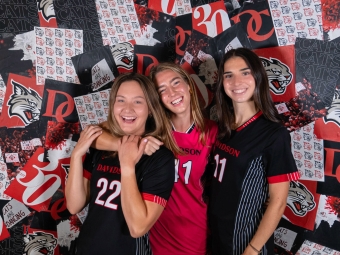 Three smiling female soccer players in Davidson College jerseys pose together in front of a red and black branded backdrop.