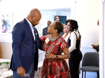 A woman in a patterned red dress talks animatedly with a man in a blue plaid suit at an indoor event.