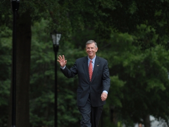 A man in a dark suit, light blue shirt, and red patterned tie smiles and waves while walking down a brick pathway lined with trees and lampposts.