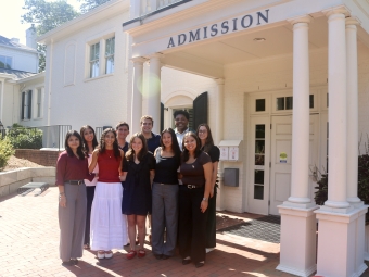 A diverse group of nine students smiling together for a group photo under the columned portico of a white "Admission" building.