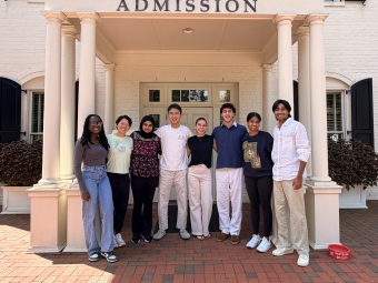 Seven diverse students stand smiling arm-in-arm in front of a white building under a porch with the word "ADMISSION" written on the pediment.