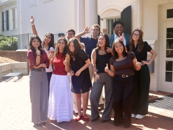 A diverse group of nine students posing playfully, smiling, and giving thumbs-up and peace signs outside a brick building.