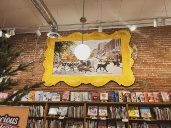A large, ornate yellow frame displaying a painting of dogs walking down a street hanging on an exposed brick wall above shelves of books.
