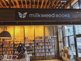 The interior of Milkweed Books featuring high wooden ceilings, tall bookshelves, and a patron browsing near a display table.