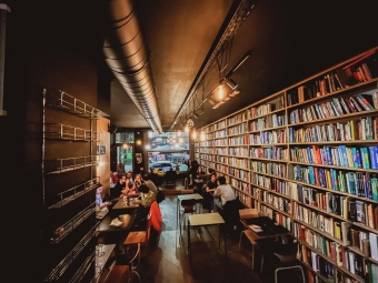 A warm, dimly lit book café featuring a long wall of filled bookshelves, seating tables, and exposed ceiling ductwork.
