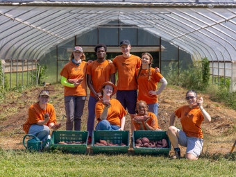 A group of students in a high tunnel, wearing matching farm shirts, in the entrance of a high-tunnel full of sweet potatoes. Three baskets of sweet potatoes are in the foreground.