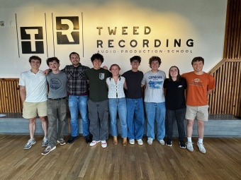 A group of eight young adults stands in a line with their arms around each other's shoulders, posing for a photo inside a modern room in front of a wall with a logo that reads "Tweed Recording Audio Production School."