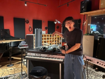A young man in glasses and a black t-shirt makes an exaggerated, surprised expression while pointing at a vintage Rhodes electric piano inside a red-walled recording studio.