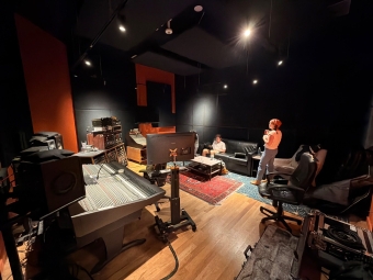 A woman stands with a cup in a dimly lit studio control room near a large mixing console, while someone else sits on a couch in the background.