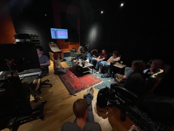Band members relax and listen in a darkened recording studio control room while seated on couches and chairs around a central coffee table.