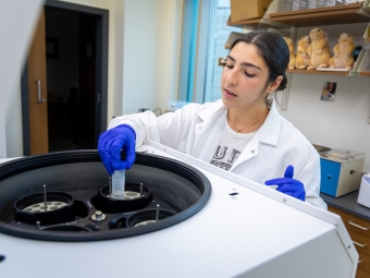 A student researcher wearing a lab coat and blue gloves carefully places a test tube into a large laboratory centrifuge.