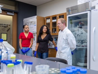 Dr. Thurtle-Schmidt, wearing a white lab coat, speaks with two student researchers wearing protective blue gloves inside a science laboratory.