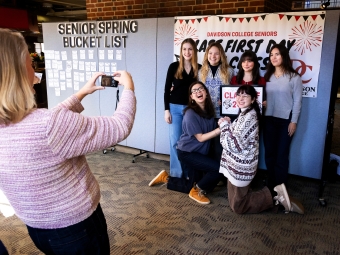 A woman takes a photo on her phone of six students posing smiling together in front of a "Senior Spring Bucket List" board and a "Class First Day" sign.
