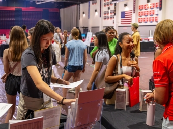 Students browse informational materials at tables during an indoor orientation fair.