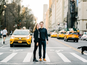 A young couple dressed in black in a crosswalk in New York City, yellow cabs visible behind them