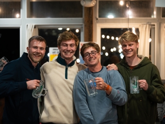 Four smiling young men stand together indoors while holding up clear glass mugs.