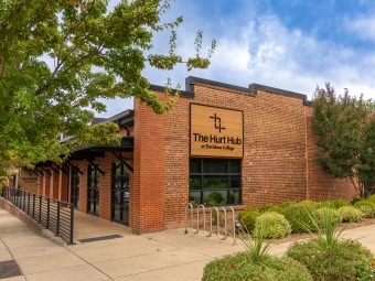 Exterior of a red brick building with a prominent sign reading "The Hurt Hub at Davidson College," surrounded by a sidewalk, bike rack, and green foliage under a partly cloudy sky.