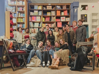 A diverse group of smiling young adults poses together inside a cozy bookstore with tall shelves filled with children's books and illustrations in the background.