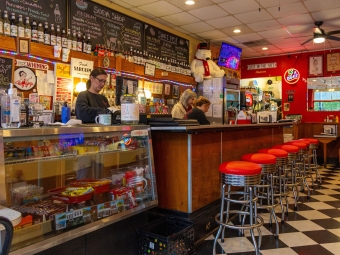 The interior of an old fashioned soda shop, with red bar stools, black and white checkerboard tiled floor, and vintage memorabilia behind the counter