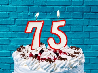 A cake in the foreground, holding candles in the shape of "75", against a blue brick wall of a retro soda shop