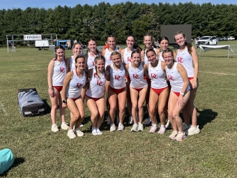 A group of female cross-country runners in matching white and red uniforms pose together on a sunny grassy field near a finish line structure.