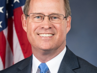 A smiling caucasian man with short dark hair, a grey suit, blue tie in front of a USA flag