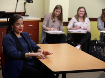 Susan Rice seated at a wooden table in a classroom. Students seated behind her are smiling