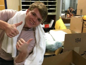 A smiling young man with a white towel draped over his shoulders gives a double thumbs-up while standing near large cardboard boxes filled with packaged items