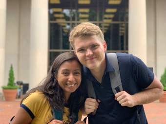 Zach and Yara smile together while wearing backpacks outdoors in front of a stone campus building with large glass windows.